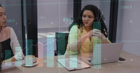 Two women reviewing overlaid financial charts at conference table, with laptop clipboard and mug