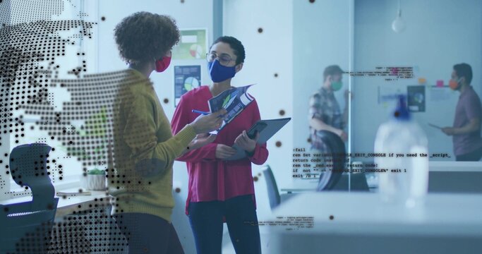 Reviewing printouts, two masked women tapping tablet screen in modern office with charts, sanitizer