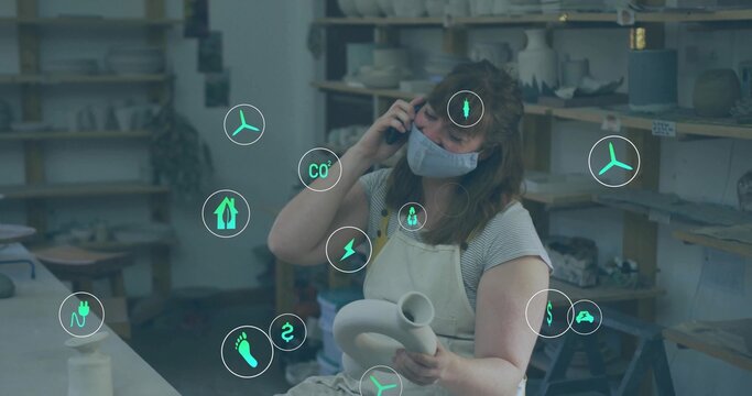 Holding clay vessel, apron-clad ceramic artist talking on smartphone at workshop, with energy icons