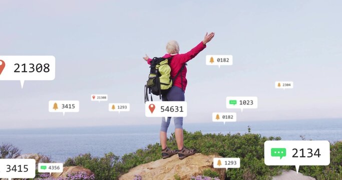 Raising arms woman wearing red jacket and boots on seaside cliff, with backpack and notifications