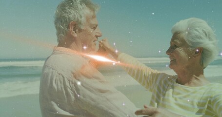 Dancing senior couple wearing casual attire and extending arms on sandy shoreline with sparkles