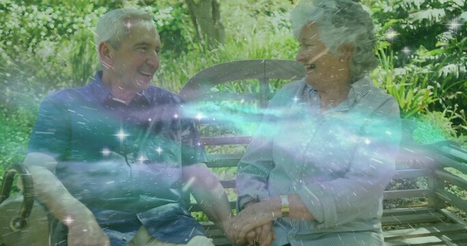 Laughing senior couple holding hands on wooden bench in garden, with wristwatch and sunlit foliage