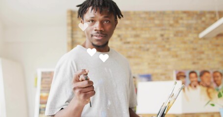 Artist with dreadlocks in paint-stained shirt pointing brush toward camera in studio, with canvases