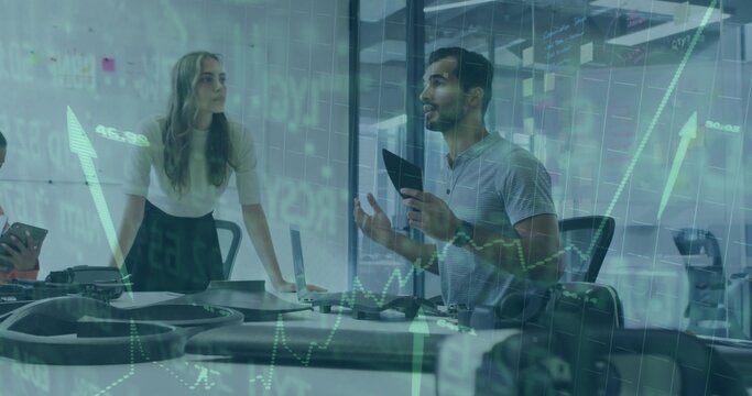 Standing man in gray polo holding tablet, gesturing in conference room with laptop, phones, graphs