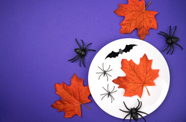 Halloween table setting with a white plate, orange autumn leaves, black spiders and bats on a purple background. Festive and spooky decoration concept for Halloween parties. Top view. Flatlay.