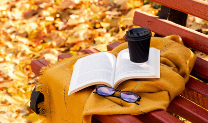 Open book, hot coffee and eyeglasses on a warm blanket outdoors on a bench in autumn park. Cozy fall atmosphere. Reading, studying, and relaxation concept. Selective focus.