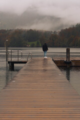 A man walks along the edge of the wooden pier, observing the autumn landscape on a rainy day