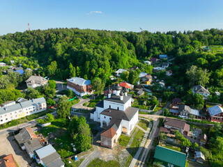 An aerial view of a small Russian Orthodox church, situated in a residential area at the base of a dense green hill with a tower visible on top.