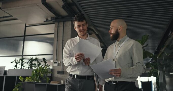 Two male coworkers walk together in a modern office holding documents and discussing work projects teamwork collaboration professional atmosphere