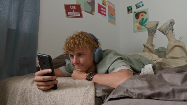 Curly haired young man using smartphone and watching funny videos online while lying on ved at home and swinging feet up in air