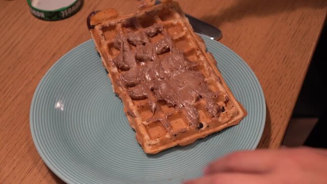A person spreading delicious chocolate hazelnut cream on a freshly baked Belgian waffle served on a blue plate for a sweet breakfast or dessert