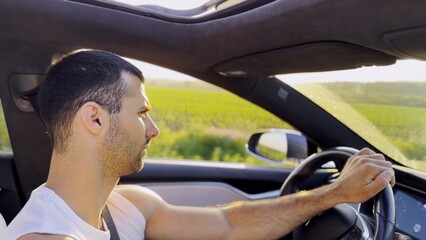 Profile of young handsome man driving a modern electric car with sunlight at background. Young male driver riding on electrical vehicle at countryside. Concept of journey and trip. Close up