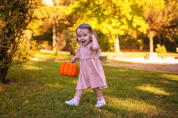 Adorable little girl, just two years old, beams with pure joy as she holds a bright orange pumpkin in an autumn park. The golden leaves and warm sunlight.