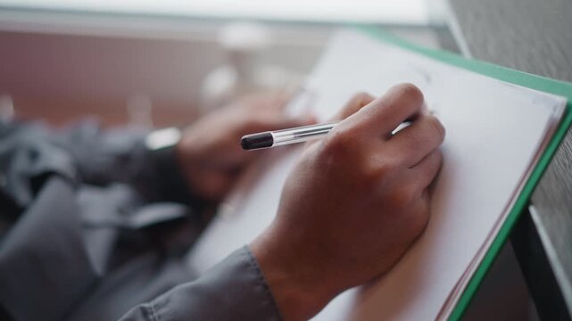 Immigrant writing with pen on white paper clipped to green folder near window, close-up of hands and focus on movement of pen, suggesting concentration, creativity, or preparation in calm indoor