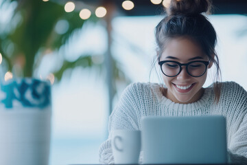 Young woman wearing glasses and a knitted sweater smiling happily while working on a laptop, enjoying remote work or studying in a modern coffee shop environment with a warm beverage