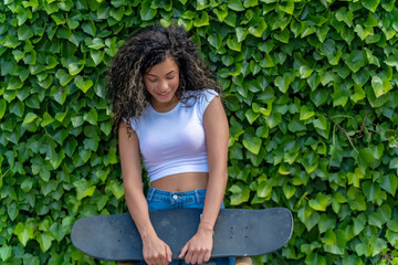 Young woman with curly hair holding a skateboard standing in front of green leaves
