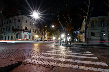 Fototapeta premium Urban street scene at nighttime featuring light trails from vehicles, pedestrian crosswalk, and illuminated buildings in a peaceful city environment.