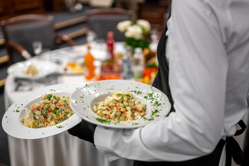 Waiter in white shirt and black apron serving two plates of Russian salad at a formal dining event with elegant table setting and floral centerpiece.