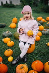 A young girl sits among pumpkins in a grassy field during autumn, happily inspecting a pumpkin