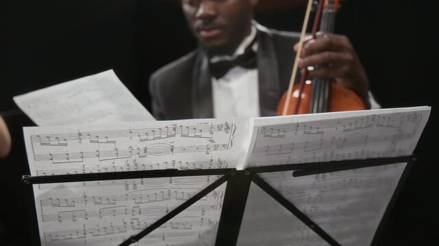 Close up shot of Black male violinist reading sheet music turning pages on note stand while rehearsing with string orchestra on stage