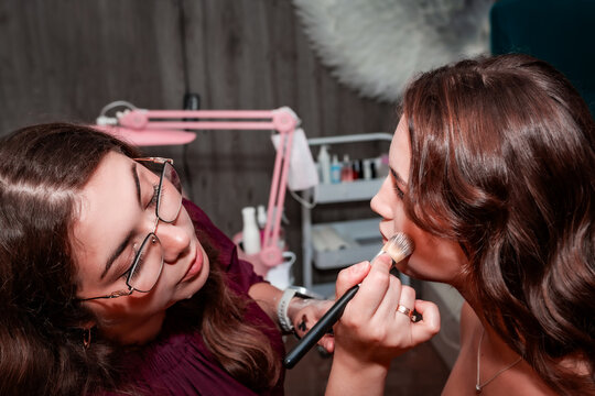 Makeup artist applying make up to a beautiful young pretty girl in a professional salon. Beauty, fashion and skin care industry. The work of a makeup artist. Blush, powder, concealer, foundation.	