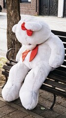A teddy polar bear with a red bow and scarf is sitting on a bench outside 