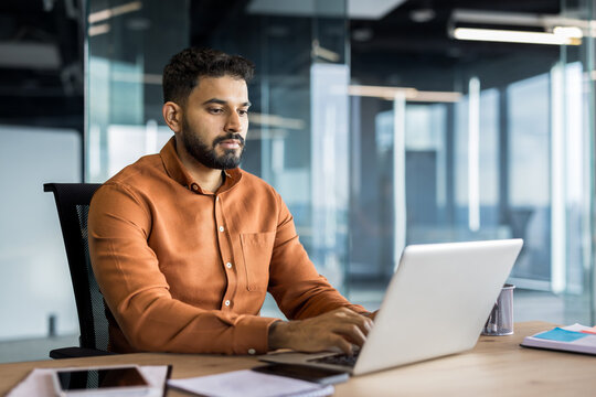Indian businessman concentrating on work, typing on a laptop at a wooden desk in a contemporary open-plan office setting, focusing on professional tasks