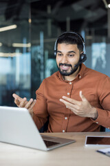 Indian man wearing headset smiling and gesturing hands while having an online video call for...