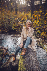 A woman in coat and jeans relaxes on a fallen tree by a serene stream in autumn