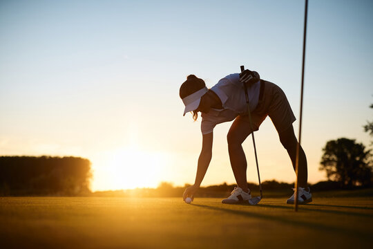 Female golfer placing golf ball on grass at sunset.