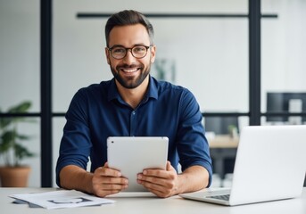 Smiling businessman in blue shirt and glasses using a tablet at his desk with laptop and papers for modern office work.