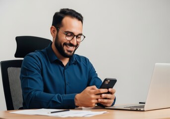 Happy smiling man wearing glasses and blue shirt using smartphone at office desk with laptop and papers