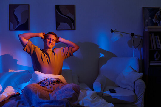 Man sitting on bed stretching arms after waking up in bedroom, smiling with eyes closed, lamp and smartphone on side table, abstract paintings on wall
