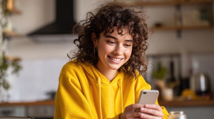 A woman with curly hair wearing a yellow hoodie smiles warmly while interacting with her smartphone in a bright kitchen during morning hours creating a cheerful atmosphere.