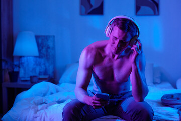 Naklejka premium Man sitting shirtless on bed listening to music with wireless headphones holding smartphone looking down illuminated by blue and purple light