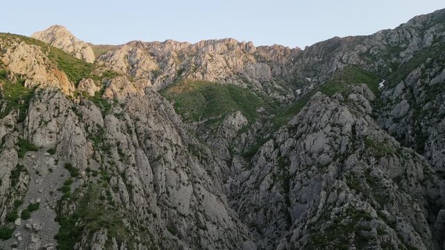 Drone tilt reveals a highland ridge above a limestone gorge in Uzbekistan with green slope, gravel ribbon and distant pass. Golden hour brightens the crest and carves shadow for a cinematic landscape