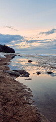 Sunrise over a tranquil coastline with icy patches and smooth stones along the sandy shore. The sky reflects in the water, creating a serene and peaceful atmosphere.