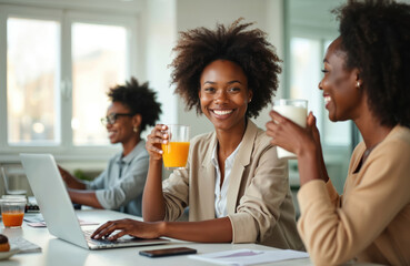 Happy African woman with curly hair enjoys orange juice at the office. A colleague laughs nearby. Healthy breakfast beverage break during work. Team communication and collaboration.