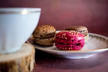 White porcelain cup with gold trim filled with dark coffee sits on a wooden coaster beside a plate of pink and brown macarons with sprinkles. Textured burgundy backdrop adds a cozy, elegant touch.