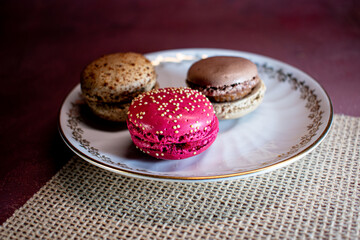 White decorative plate on burlap holds colorful macarons, creating a cozy, inviting scene. Soft textures and warm tones evoke a relaxing morning and the pleasure of sweet indulgence.