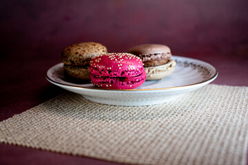 White decorative plate on burlap holds colorful macarons, creating a cozy, inviting scene. Soft textures and warm tones evoke a relaxing morning and the pleasure of sweet indulgence.