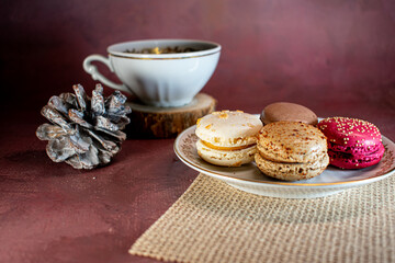 White cup of black coffee on a wooden coaster beside a pine cone and a plate of colorful macarons—white, brown, and pink—on a textured placemat. Dark background adds a warm, cozy ambiance.