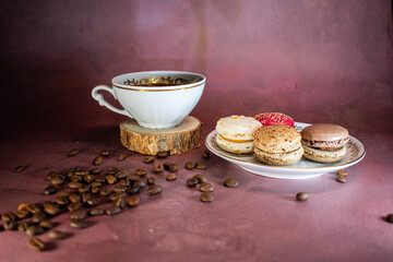 White coffee cup with gold trim on wooden coaster beside a plate of colorful macarons—chocolate, coffee, pink, and vanilla. Coffee beans scattered on a burgundy surface create a warm, inviting scene.