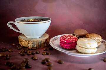 White coffee cup with gold trim on wooden coaster beside a plate of colorful macarons—chocolate, coffee, pink, and vanilla. Coffee beans scattered on a burgundy surface create a warm, inviting scene.