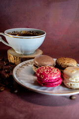 White coffee cup with gold trim on wooden coaster beside a plate of colorful macarons—chocolate, coffee, pink, and vanilla. Coffee beans scattered on a burgundy surface create a warm, inviting scene.
