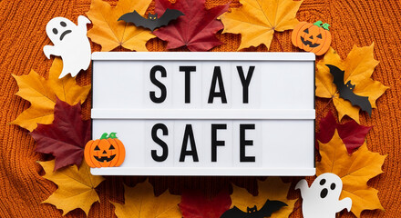Lightbox with "Stay Safe" message, surrounded by autumn leaves, pumpkins, and ghosts on orange knit fabric Halloween safety concept