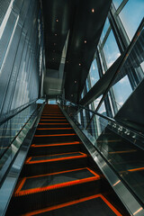 A low-angle shot of a modern escalator with glowing orange steps, sleek metal railings, and a reflective ceiling, surrounded by large glass windows