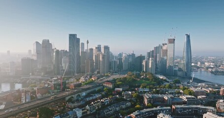 Australia, Sydney: Aerial view of Sydney Harbour Bridge and city skylines skyscrapers reflecting in water at misty sunrise, Barangaroo Reserve and wharves in foreground. Drone flight footage panorama - Powered by Adobe