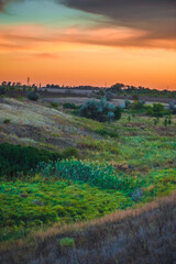 Lush green and yellow grass in a rolling field under a striking orange and purple evening sky, capturing the last light of a summer day.