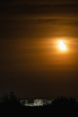 A golden-orange half moon over a cityscape at dusk, with buildings and trees silhouetted against a cloudy sky.